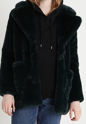 Veste en fausse fourrure noire avec grand col portée sur un sweat à capuche noir et un jean bleu sur une personne debout.