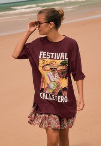 Camiseta amplia color granate con gráfico frontal "FESTIVAL CALLETERO" e ilustración colorida. Combinada con una falda con estampado y volantes.