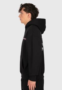Profil d'un jeune homme avec des cheveux bouclés portant un sweatshirt à capuche noir avec un texte rouge et blanc, se tenant devant un fond gris clair uni.