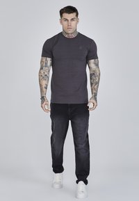 Donkergrijs aansluitend T-shirt met korte mouwen, ronde hals, geborduurd logo op de borst, gedragen met zwarte jeans en witte sneakers.