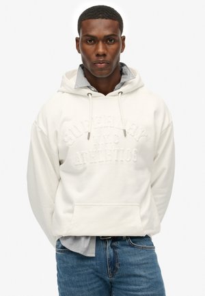 White fleece hoodie with front pocket, embroidered logo, and drawstring hood, layered over a light blue striped shirt and jeans.