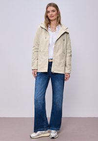 Mujer con chaqueta beige con capucha sobre camisa blanca, jeans anchos azules y zapatillas plateadas, de pie contra un fondo claro y sencillo.