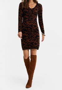 Femme portant une robe à manches longues noire et marron à motifs, assortie à de grandes bottes en daim marron et des bracelets au poignet droit.