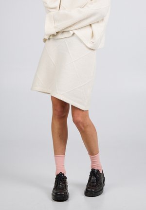 Unterkörper in einem cremefarbenen, strukturierten Rock, hellrosa Socken und schwarzen, geschnürten Lederschuhen vor einem schlichten, hellen Hintergrund.