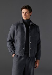 Szara wełniana kurtka z zapięciem na guziki z przodu, kołnierzem typu rozłożysty oraz elastycznymi mankietami. Noszona na dopasowanym ciemnym golfie i eleganckich spodniach.