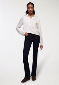 Camicia bianca a maniche lunghe con bottoni e tasca, abbinata a jeans slim-fit blu scuro e stivali alla caviglia marroni, in piedi contro uno sfondo chiaro.
