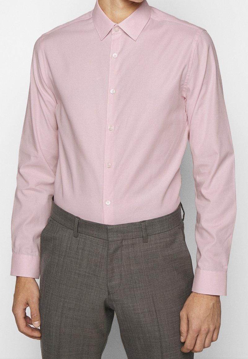 Camicia a maniche lunghe rosa chiaro con colletto a punta, caratterizzata da una chiusura con bottoni sul davanti, una texture liscia e una lucentezza sottile, abbinata a pantaloni grigi a quadri.