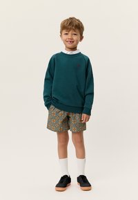 Τιρκουάζ φούτερ με ριμπ μανσέτες, σορτς με μοτίβο σε πορτοκαλί και μπλε αποχρώσεις, λευκές κάλτσες μέχρι τον αστράγαλο και μαύρα αθλητικά παπούτσια. Κοντά καστανά μαλλιά.