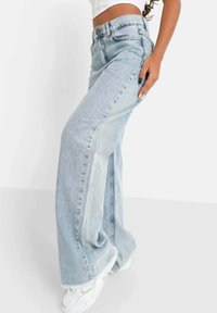 Donna che indossa jeans a gamba larga blu chiaro e sneakers bianche, in posa con una mano sul fianco contro uno sfondo neutro.