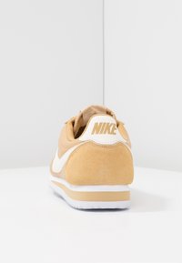 Nike-sneaker i ljust tanmocka, med en vit swoosh, stoppad krage och en strukturerad gummisula. Diagonala linjer betonar designen.