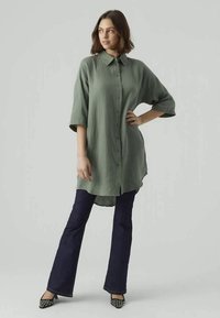 Camisa texturizada verde-oliva, com colarinho e mangas até ao cotovelo, combinada com jeans azul-escuro de boca de sino e calçado com padrão.