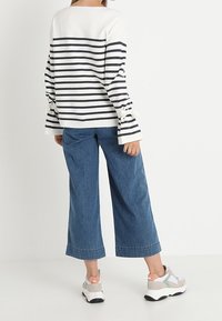 Persona che indossa una maglietta a maniche lunghe bianca e nera a righe con polsini annodati, jeans blu a gamba larga e cropped, e sneakers bianche con accenti pastello.