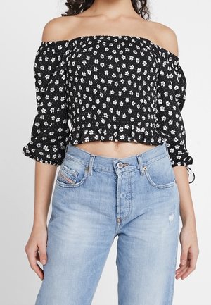 Žena v černém crop topu s odhalenými rameny a bílým květinovým vzorem, světle modrých džínách s vysokým pasem.