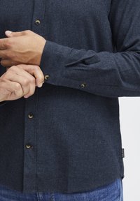 Camisa de franela azul oscuro con collar abotonado, botones marrones y una textura tejida. Incluye una pequeña etiqueta de logo cerca del dobladillo.