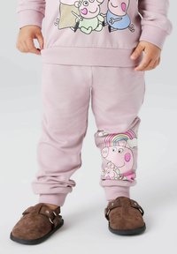 Gaiši rozā džemperis ar multfilmu tēliem, kombinēts ar rozā biksēm ar cūkas dizainu un brūnām zamšādas slip-on kurpēm.