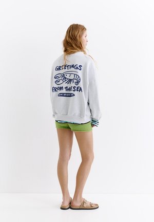 Jeune femme debout, tournée vers la droite, portant un sweat-shirt gris clair avec un homard bleu et le texte « Greetings from the sea », un short vert et des sandales.