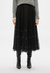 Μαύρη midi φούστα από δαντέλα με μαργαριταρένιες λεπτομέρειες, σχεδιασμένη με στρώσεις και απαλό, διαφανές ύφασμα. Συνδυάζεται με εφαρμοστά μαύρα δερμάτινα μπότες.