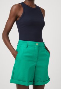 Marineblaue, gerippte ärmellose Bluse kombiniert mit leuchtend grünen, taillierten Shorts mit Knopfverschluss und umgeschlagenen Säumen, gefertigt aus einem glatten Stoff.