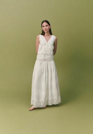 Femme debout devant un fond vert olive, portant une robe blanche sans manches avec des détails en dentelle et des colliers superposés, les mains derrière le dos.