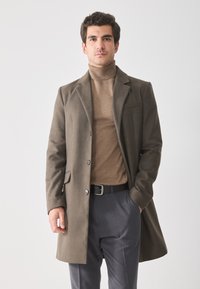 Hombre con suéter de cuello alto marrón, abrigo verde oliva, pantalones grises y cinturón negro, de pie con una mano en el bolsillo contra un fondo blanco.