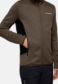 Braune Zip-Jacke aus strukturiertem Material, mit schwarzen Seitenpaneelen und Reißverschlusstaschen. Sie hat einen hohen Kragen und ein Logo auf der Brust.