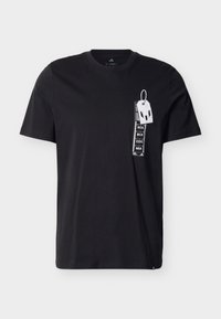 Μαύρο βαμβακερό μπλουζάκι με λευκό γραφικό και κείμενο, καθώς και σχεδίαση ετικέτας στην αριστερή πλευρά του στήθους. Κοντά μανίκια και στρογγυλή λαιμόκοψη.