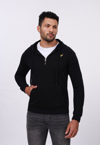 Schwarze Zip-Hoodie mit einer Vordertasche, die ein kleines goldenes Logo auf der linken Brust trägt, über einem weißen T-Shirt getragen. Glatte Stofftextur.