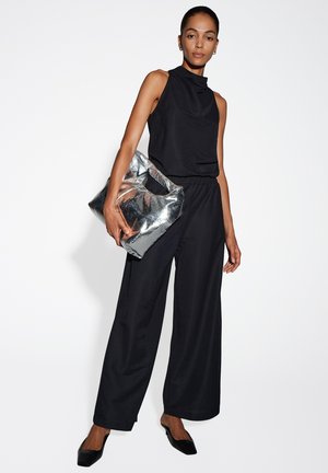 Combinaison noire sans manches avec un col montant, coupe décontractée et jambes larges ; assortie à un sac à main argenté brillant et des chaussures noires à talons pointus.