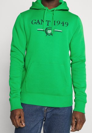 Zielona bluza z kapturem z przednią kieszenią, nadrukiem "GANT 1949" i logo, noszona przez osobę o ciemnej karnacji w niebieskich dżinsach.