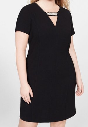 Kobieta ubrana w czarną sukienkę z krótkim rękawem i dekoltem w kształcie litery V, ozdobioną poziomym paskiem z perełkami na wysokości klatki piersiowej.