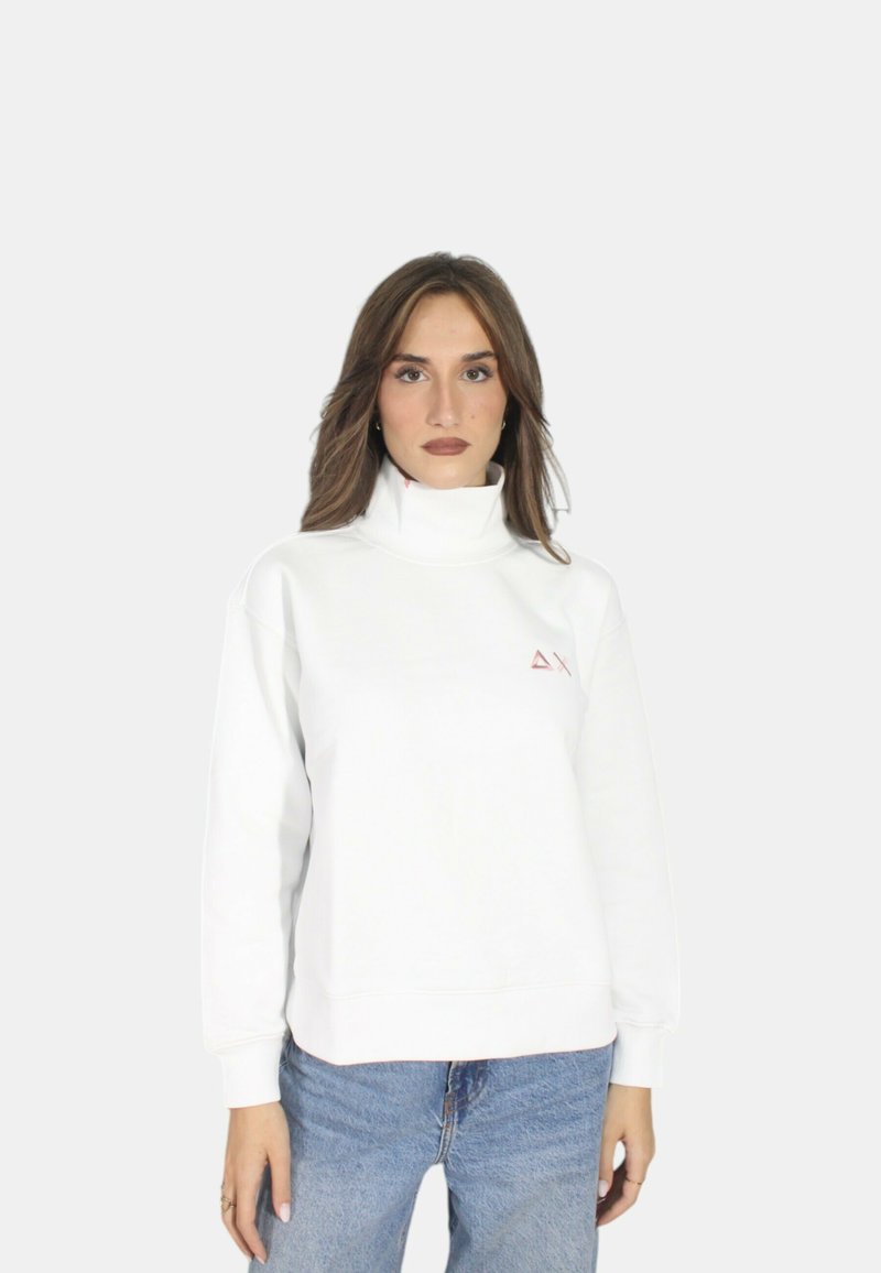 Felpa bianca con colletto alto, polsini a coste e un piccolo logo triangolare rosso sul petto. Abbinata a jeans blu chiaro.