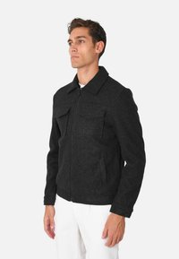 Giacca nera in misto lana con zip frontale, colletto a punta, due taschini sul petto e tasche laterali; vestibilità classica con maniche lunghe e tessuto testurizzato.