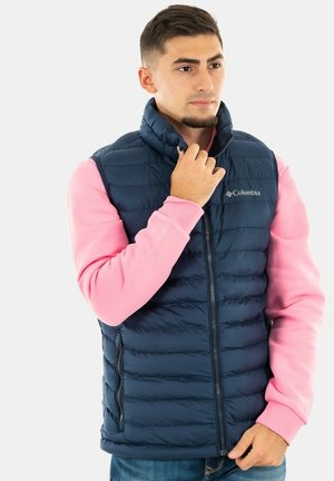 Chaleco acolchado azul marino con cuello alto y cremallera frontal. Presenta acolchado horizontal y un logo en el lado izquierdo del pecho, llevado sobre una sudadera rosa.