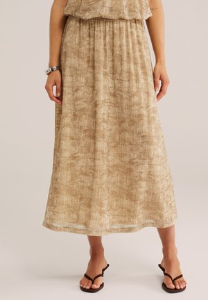 Femme portant une jupe midi beige à texture avec une taille élastique, associée à des sandales noires à lanières, debout sur un fond clair.