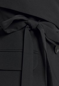 Vêtement en tissu noir avec une ceinture nouée à la taille, une texture lisse, et une poche latérale avec un détail de couture horizontale.