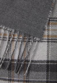 Grå uldscarf med frynser ligger på en beige, brun og sort plaid-mønstret uldstof.