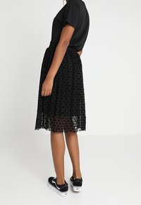 Musta t-paita yhdistetty polvimittaisen, läpikuultavan mustan pilkullisen hameen kanssa. Hameessa on mukava joustava vyötärönauha, ja asu on yhdistetty mustiin lenkkareihin.