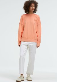 Peach-colored sweatshirt with a relaxed fit, crew neck, and ribbed cuffs. Paired with white pants and light-colored sneakers.