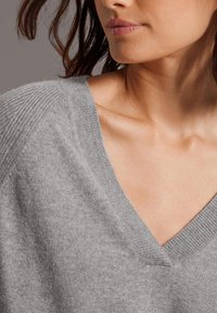 Frau, die einen weichen grauen V-Ausschnitt-Pullover trägt, der ihr Schlüsselbein sowie einen Teil ihrer Schulter und ihres Nackens zeigt, vor einem schlichten Hintergrund.