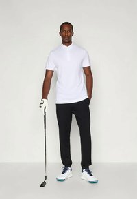 Άσπρο πόλο μπλουζάκι με κοντό γιακά, συνδυασμένο με μαύρο παντελόνι. Φορά γάντι γκολφ, κρατά γκολφ κλαμπ και άσπρα αθλητικά παπούτσια με μπλε λεπτομέρειες.