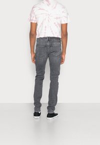 Persona con jeans gris ajustados, camiseta de rayas blanca y rosa clara, y zapatillas Converse negras, de pie contra una pared blanca lisa.