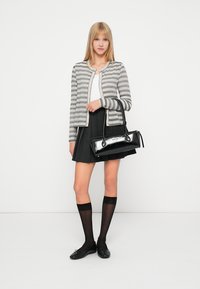 Ung kvinne iført stripete kardigan, svart plissert skjørt, knelange gjennomsiktige sokker, svarte leiligheter, som holder en svart håndveske mot en hvit bakgrunn.