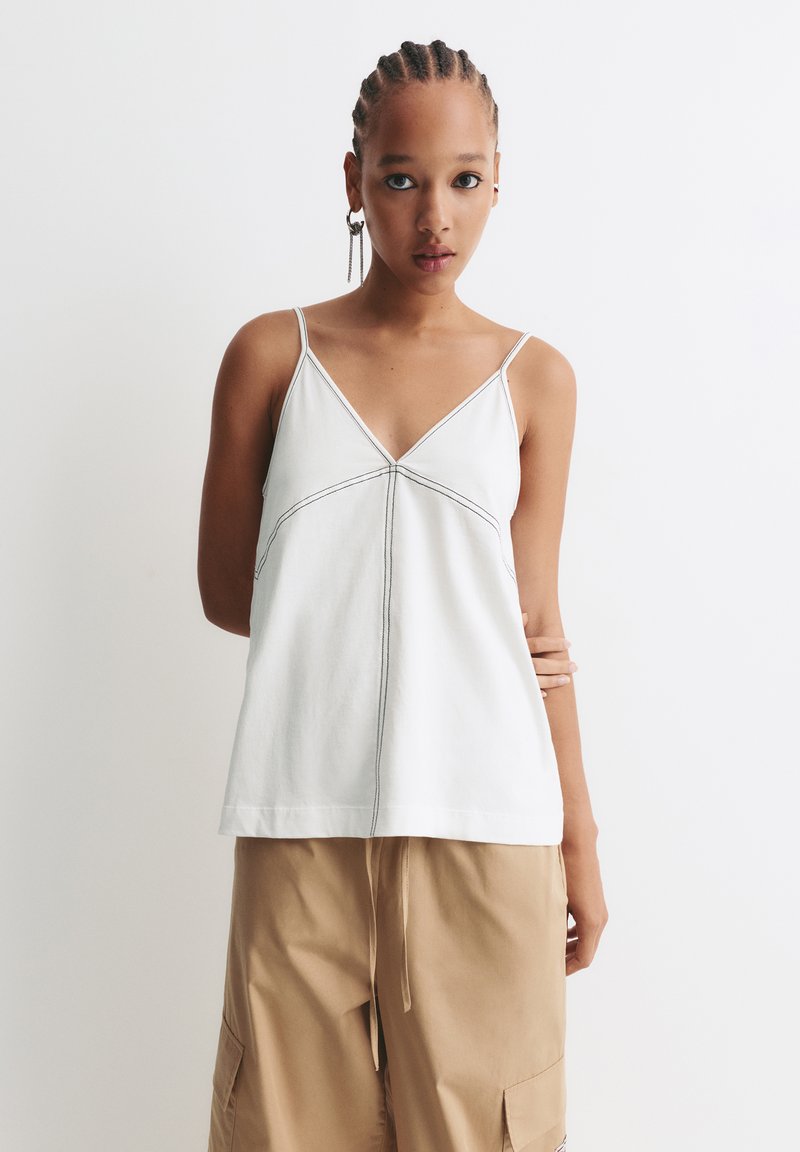 Jonge vrouw met gevlochten haar, gekleed in een witte mouwloze top met zwarte stiksels en beigekleurige cargobroek, staande tegen een witte achtergrond.