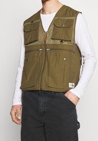 Man som bär en olivgrön mångficksväst över en vit långärmad skjorta och mörka byxor, med en hand i fickan, stående mot en enkel bakgrund.