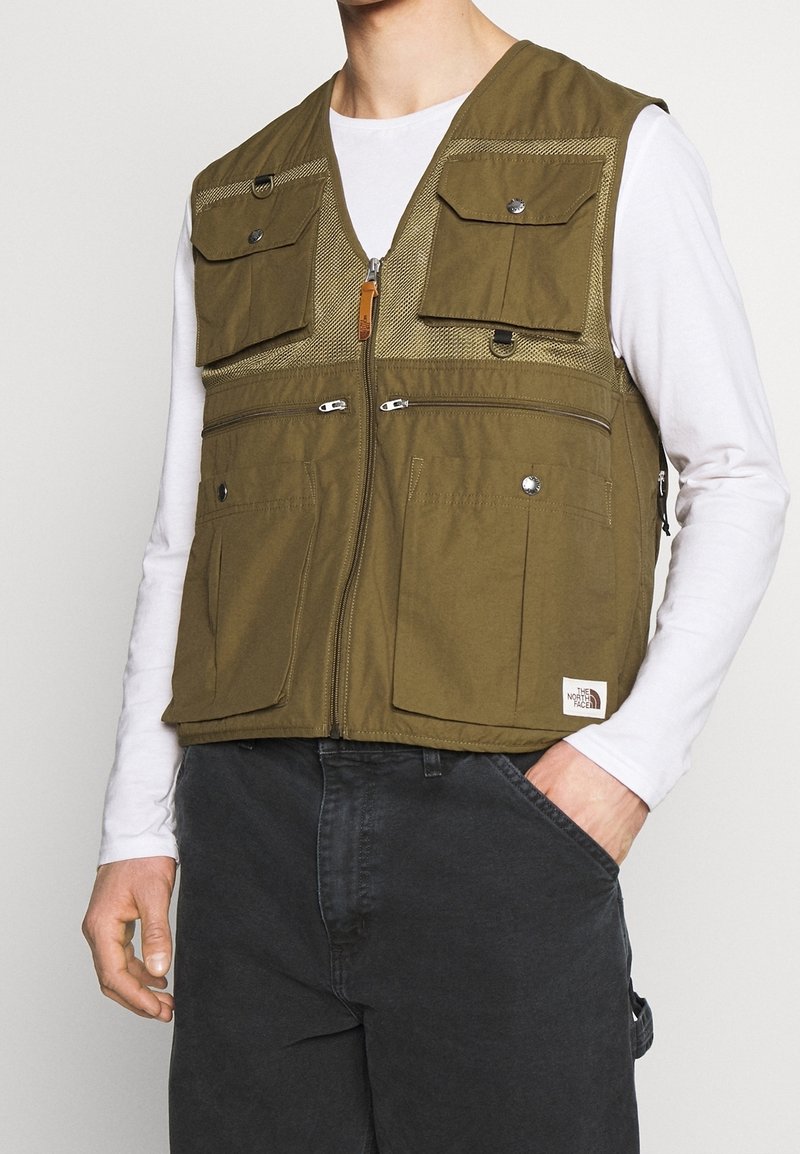 Man som bär en olivgrön mångficksväst över en vit långärmad skjorta och mörka byxor, med en hand i fickan, stående mot en enkel bakgrund.