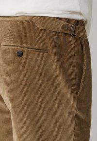 Bruine corduroy broek heeft verticale ribbels, een platte voorkant, riemlussen, een zijzak en een enkele achterzak met een knoop.