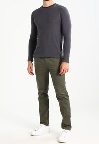 Maglia grigia scura a maniche lunghe con tasca, abbinata a pantaloni verde oliva e sneakers bianche, che mostrano un design atletico semplice.