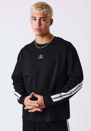 Joven con el pelo corto y decolorado, lleva una camisa negra de manga larga con rayas blancas en las mangas y un collar de cadena plateada, manos entrelazadas.