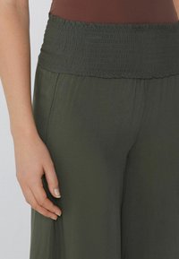 Olivově zelené kalhoty s širokým nařaseným pasem, vyrobené z hladkého materiálu a s uvolněným střihem. Detail ruky položené na boku.