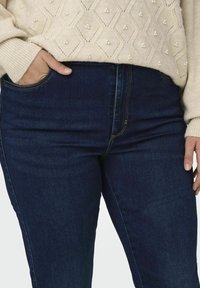 Ljus beige tröja med texturerade diamantmönster och små dekorationer, ihop med mörkblå denimjeans med sidofickor.